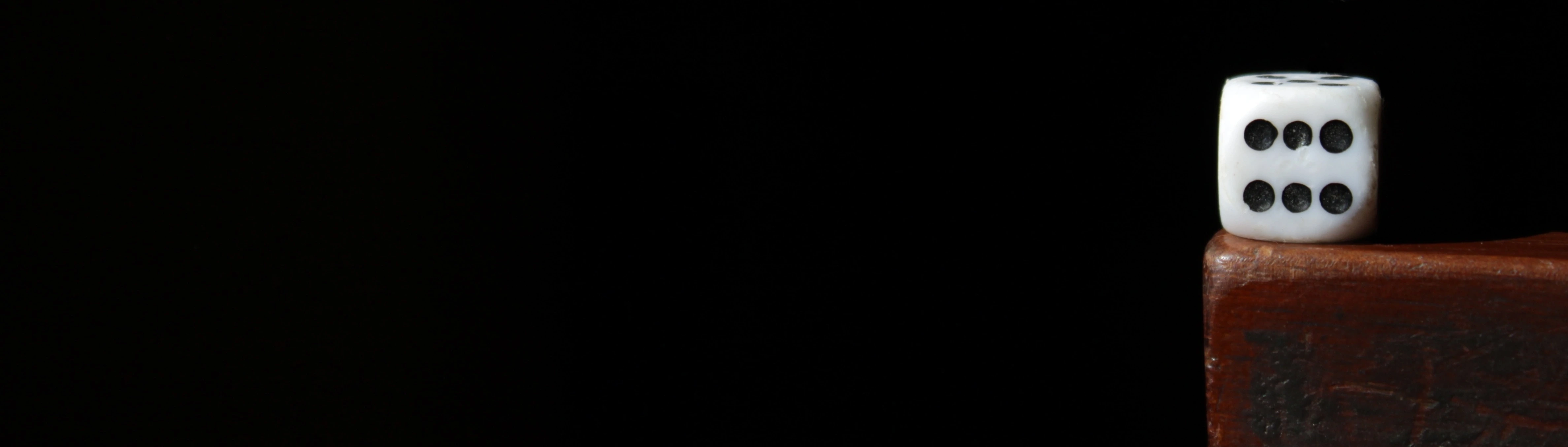 A close up of the side of a dice, showing the number 6. The dice is placed at the corner of a brown, wooden table edge.This portrays small, but with a big impact, and reflects